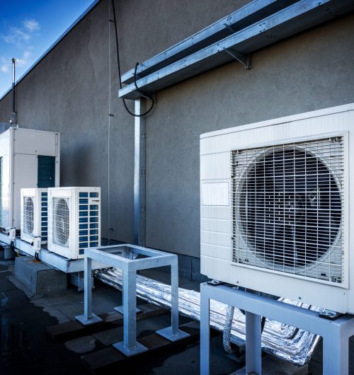 Square air-conditioning unit on the roof with a round fan. In the background gradually receding other units that are out of focus. On the right side light blue sky and commercial space.